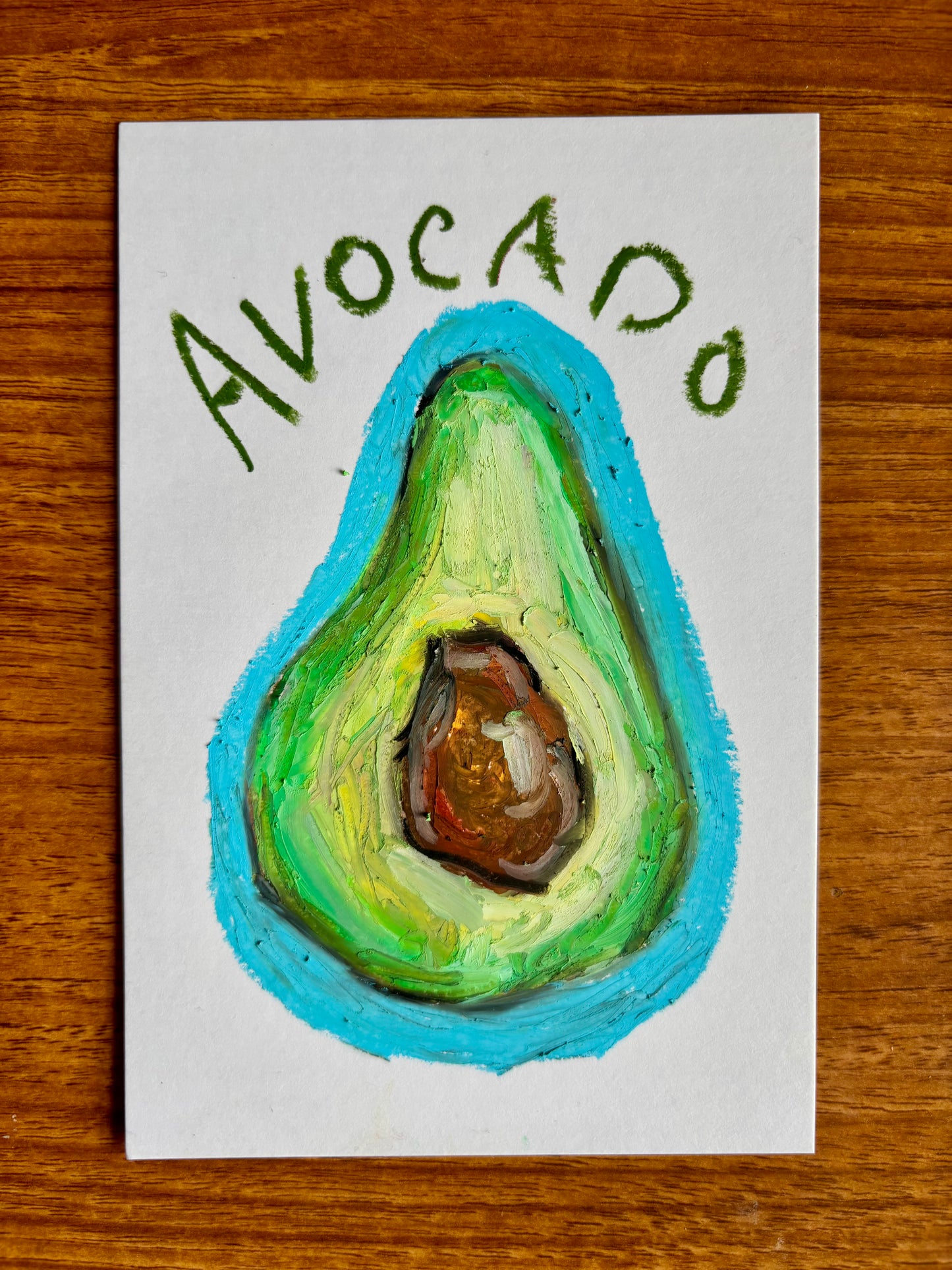 Small art print of a hand drawn avocado with green and brown tones with a bright blue background, with the text "Avocado" displayed on a wooden table background