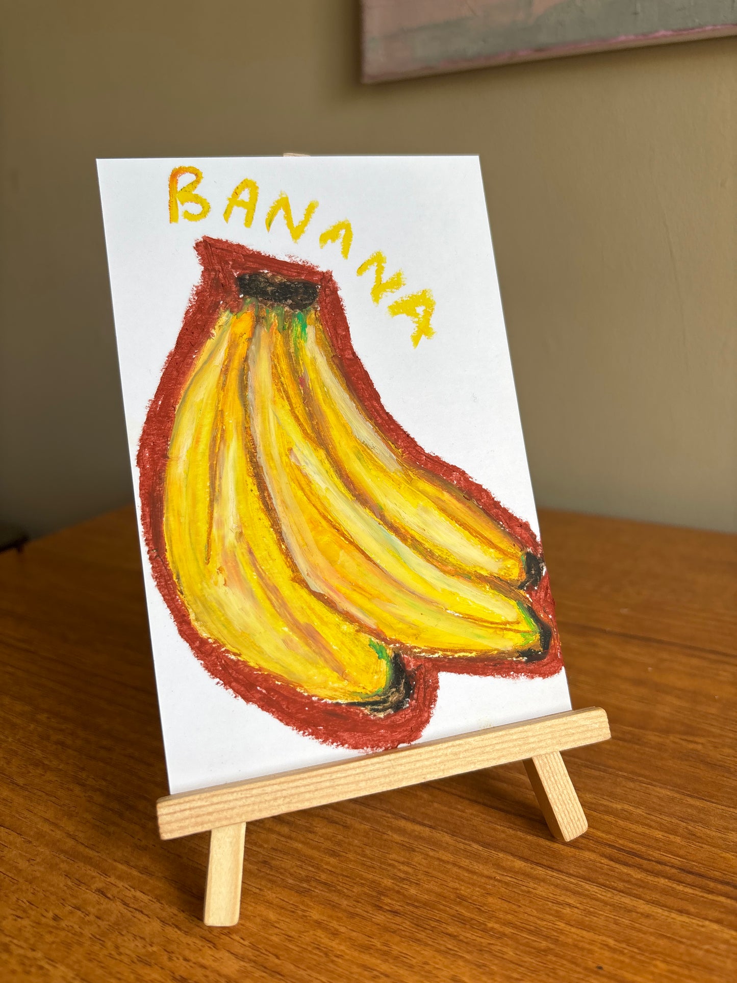 A small white drawing on a wooden easel, depicting a bunch of yellow bananas with a red outline and the word 'BANANA' written in yellow at the top, set on a wooden table.