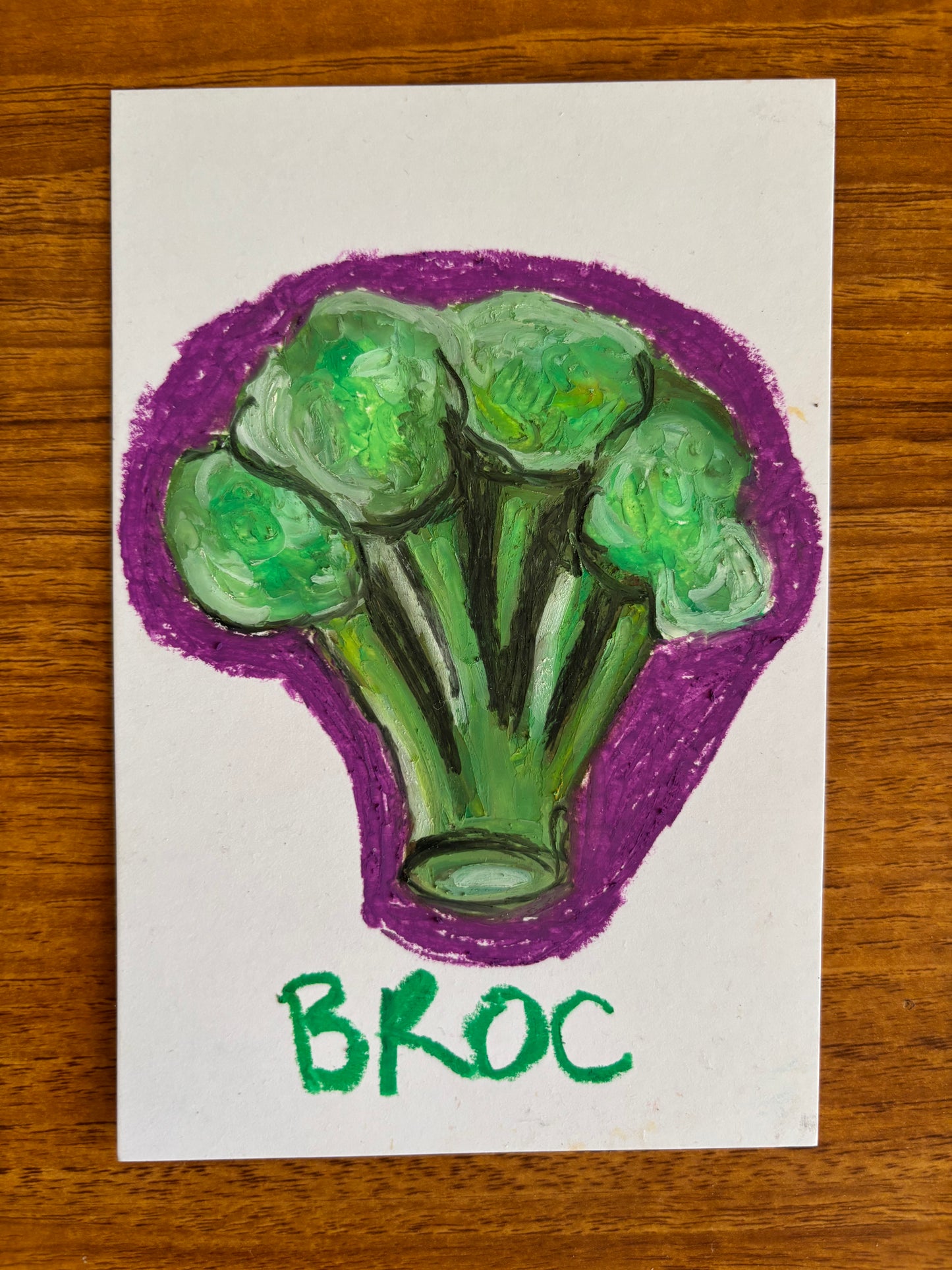 A hand-drawn illustration of a broccoli floret on a white card, placed on a wooden surface. The drawing features green broccoli with a purple outline, and the word 'BROC' written below in green.