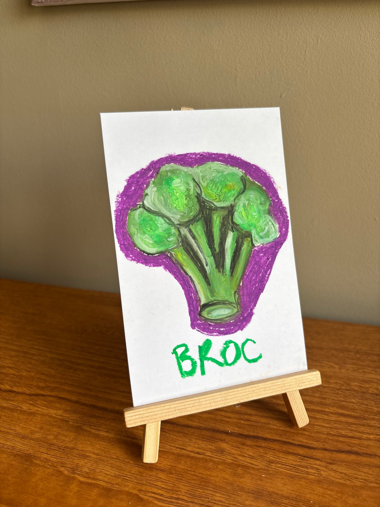 Broccolli floret hand drawn with oil pastels with a purple outline, on small card displayed on a small wooden easel
