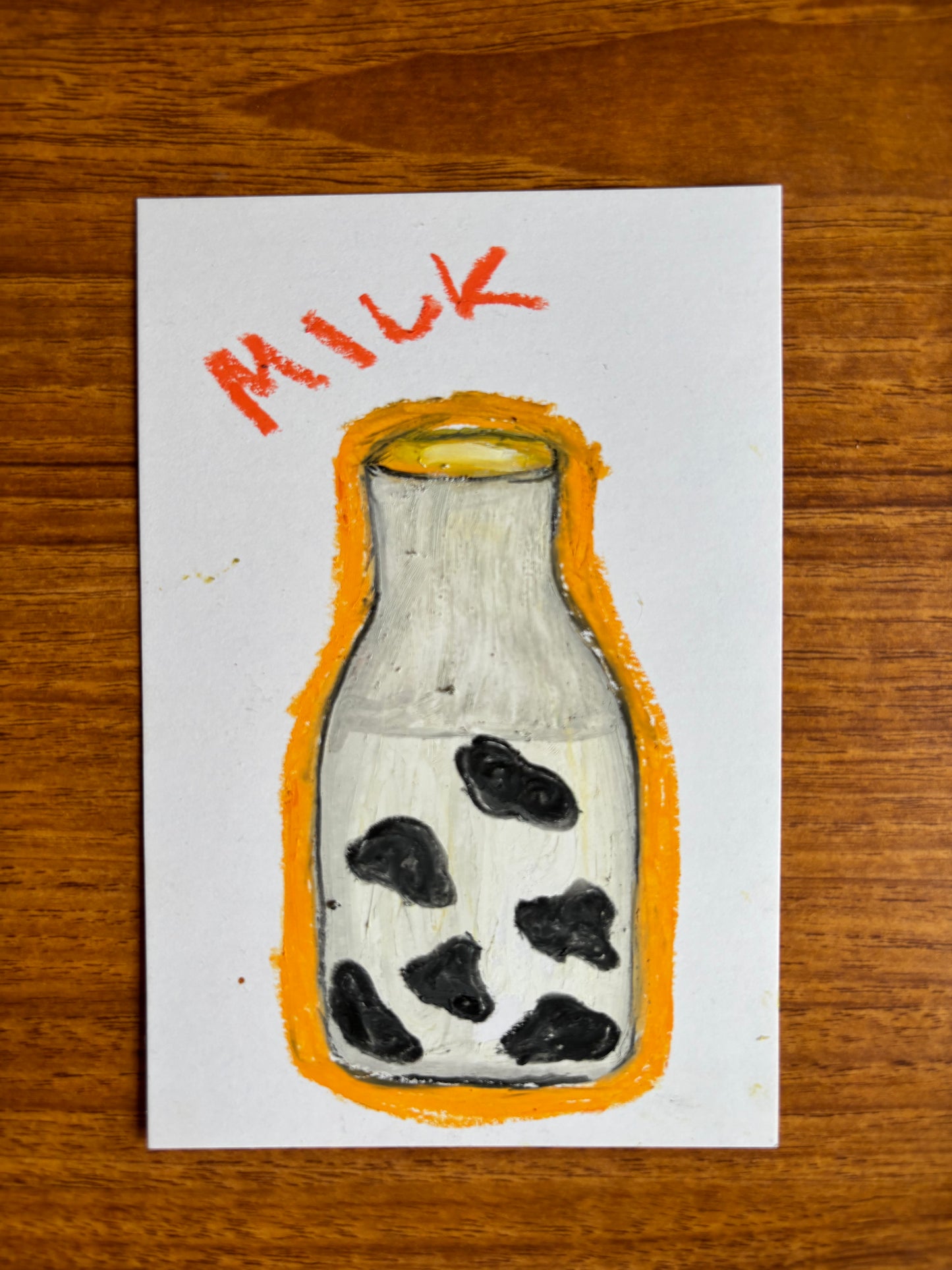 A square white art print or card featuring a hand-drawn illustration of a milk bottle with black spots, an orange outline, and the word 'MILK' in orange at the top, placed on a wooden surface.