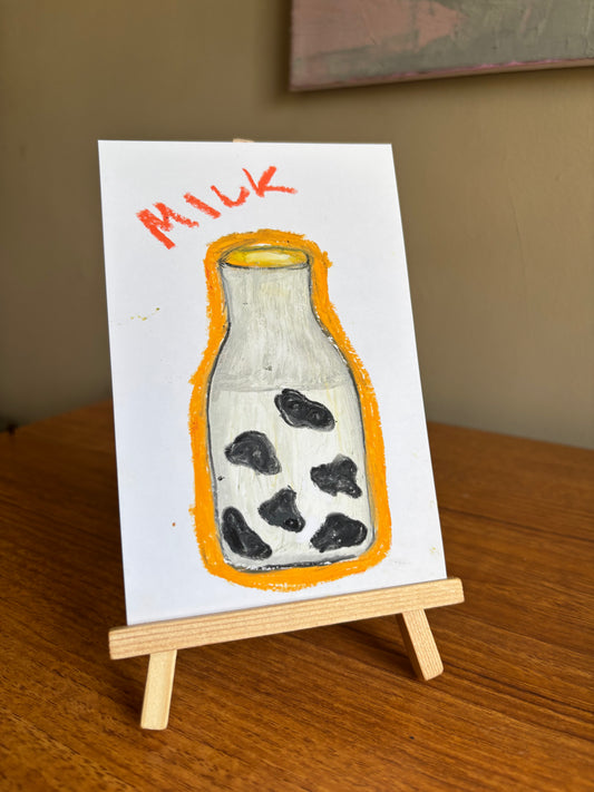 A small hand-painted artwork on a white card, depicting a milk bottle with black spots inside, an orange outline, and the word 'MILK' in orange at the top. The card is displayed on a miniature wooden easel on a wooden table.
