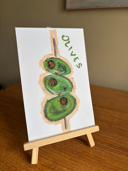 A small white card painted with three green olives on a wooden skewer and the word “OLIVES” in green, displayed on a small wooden easel on a wooden table.