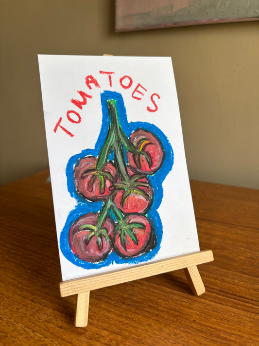 A small hand-painted canvas showing a cluster of red tomatoes on a vine with the word 'TOMATOES' written above, displayed on a miniature wooden easel on a wooden table.