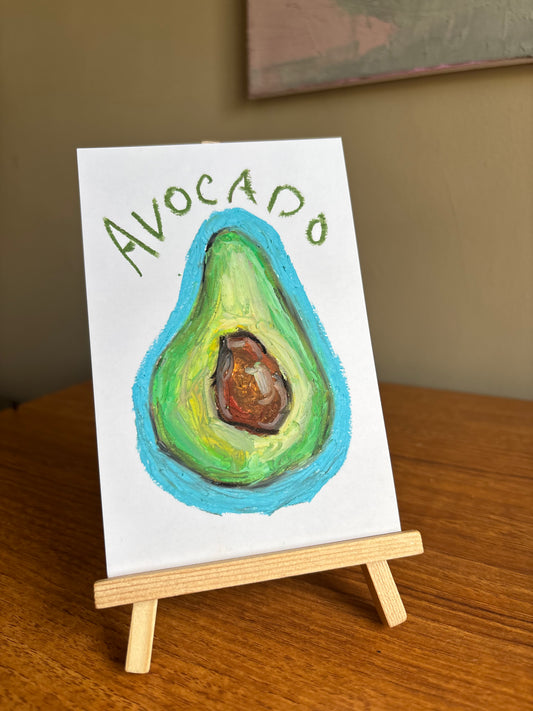 A small hand-painted canvas featuring an avocado illustration with the word 'AVOCADO' written above it, displayed on a miniature wooden easel on a wooden table.