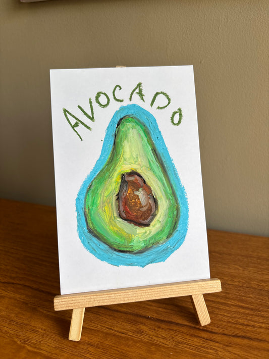 A small white art print or card on a wooden easel, featuring a painted avocado with green and brown tones, a bright blue outline, and the word “AVOCADO” written above in green. The easel sits on a wooden table against a beige wall.