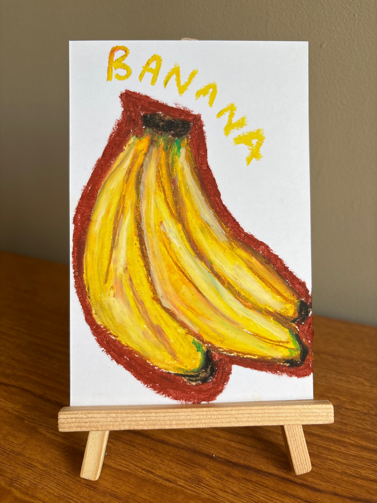 A small white art print or card showing a painted bunch of yellow bananas outlined in reddish brown, with the word "BANANA" in yellow at the top, displayed on a wooden easel on a wooden table against a beige wall.