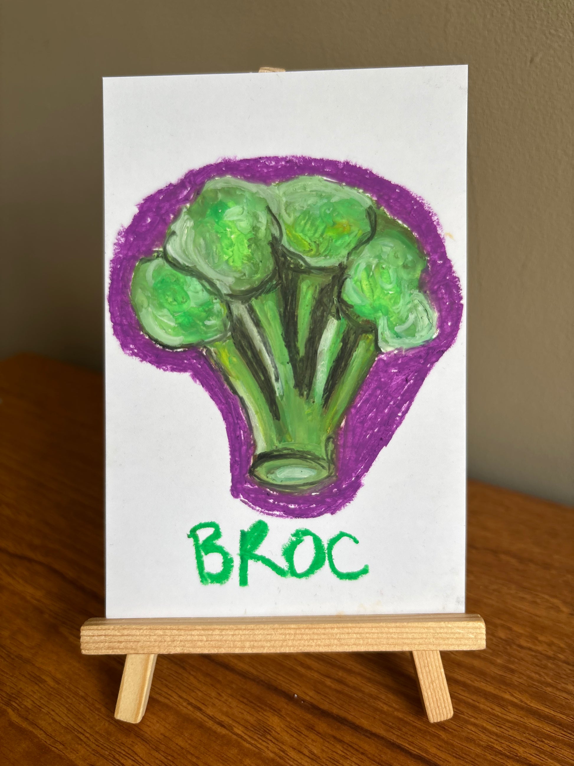 Oil pastel drawing of a broccoli floret with a purple outline and the text "Broc" on card displayed on a small wooden easel 