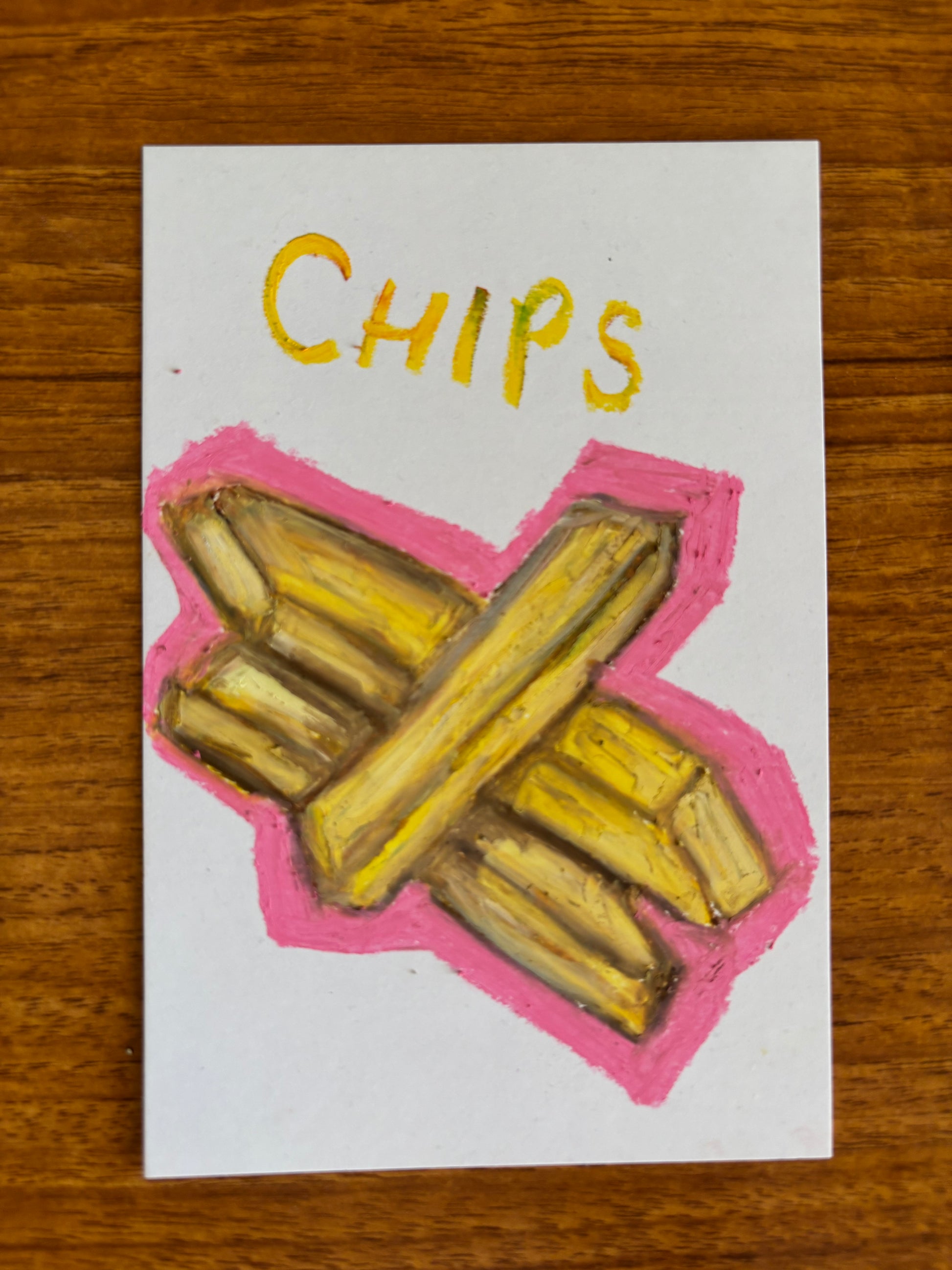 A small white art card featuring a hand-drawn illustration of french fries in yellow with a pink outline, and the word 'CHIPS' written in yellow at the top, placed on a wooden surface.