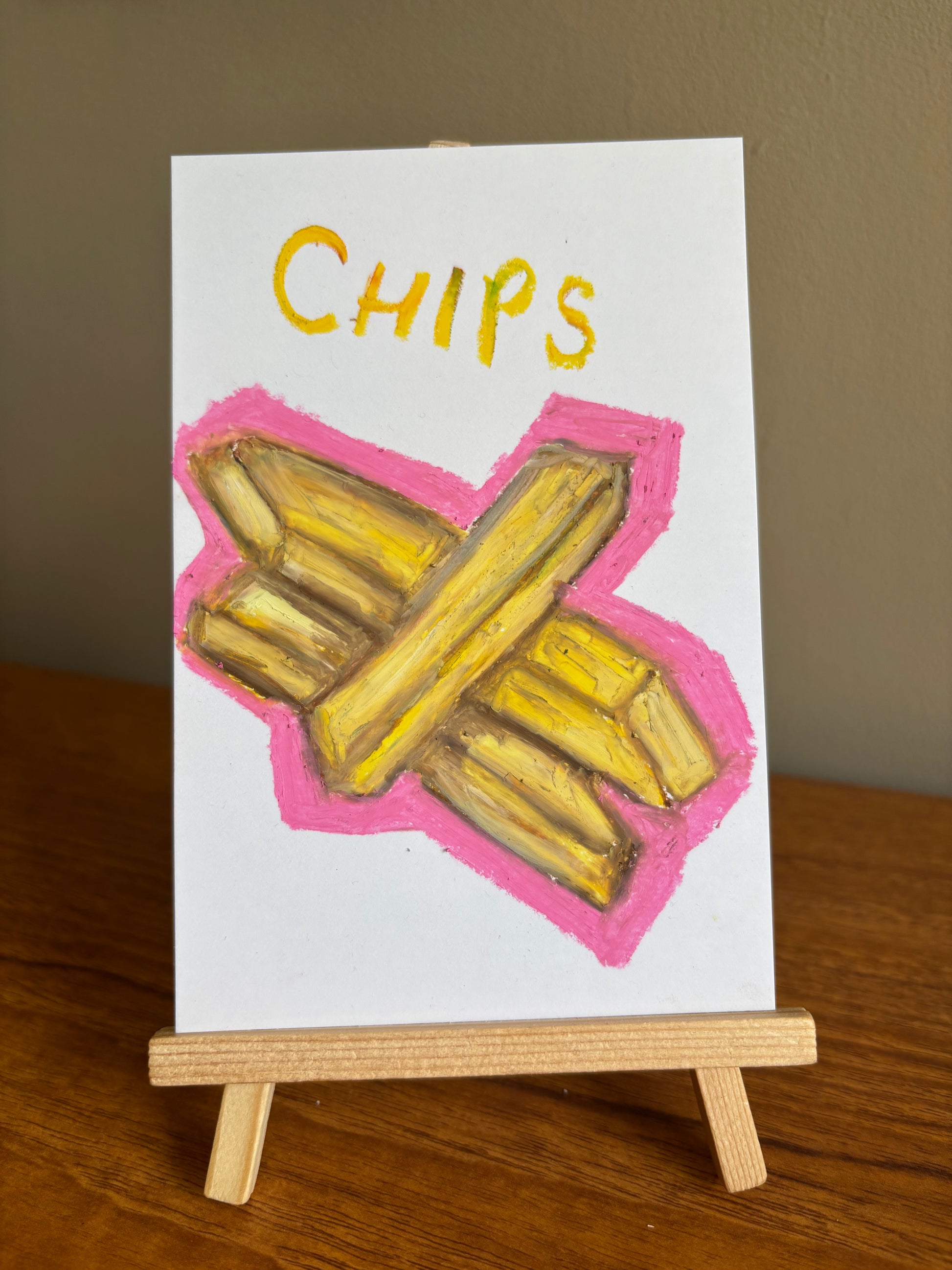 A small white art print or card on a wooden easel, featuring a hand-drawn illustration of yellow chips crossed over each other with a pink outline, and the word "CHIPS" in yellow at the top.