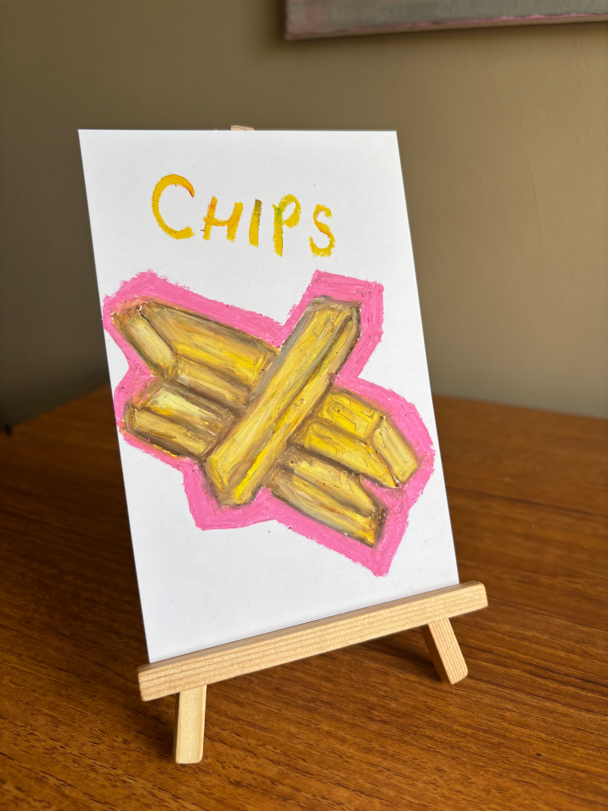 A small hand-painted artwork on white paper showing yellow french fries crossed over each other with a pink outline and the word "CHIPS" in yellow at the top, displayed on a miniature wooden easel on a wooden table.