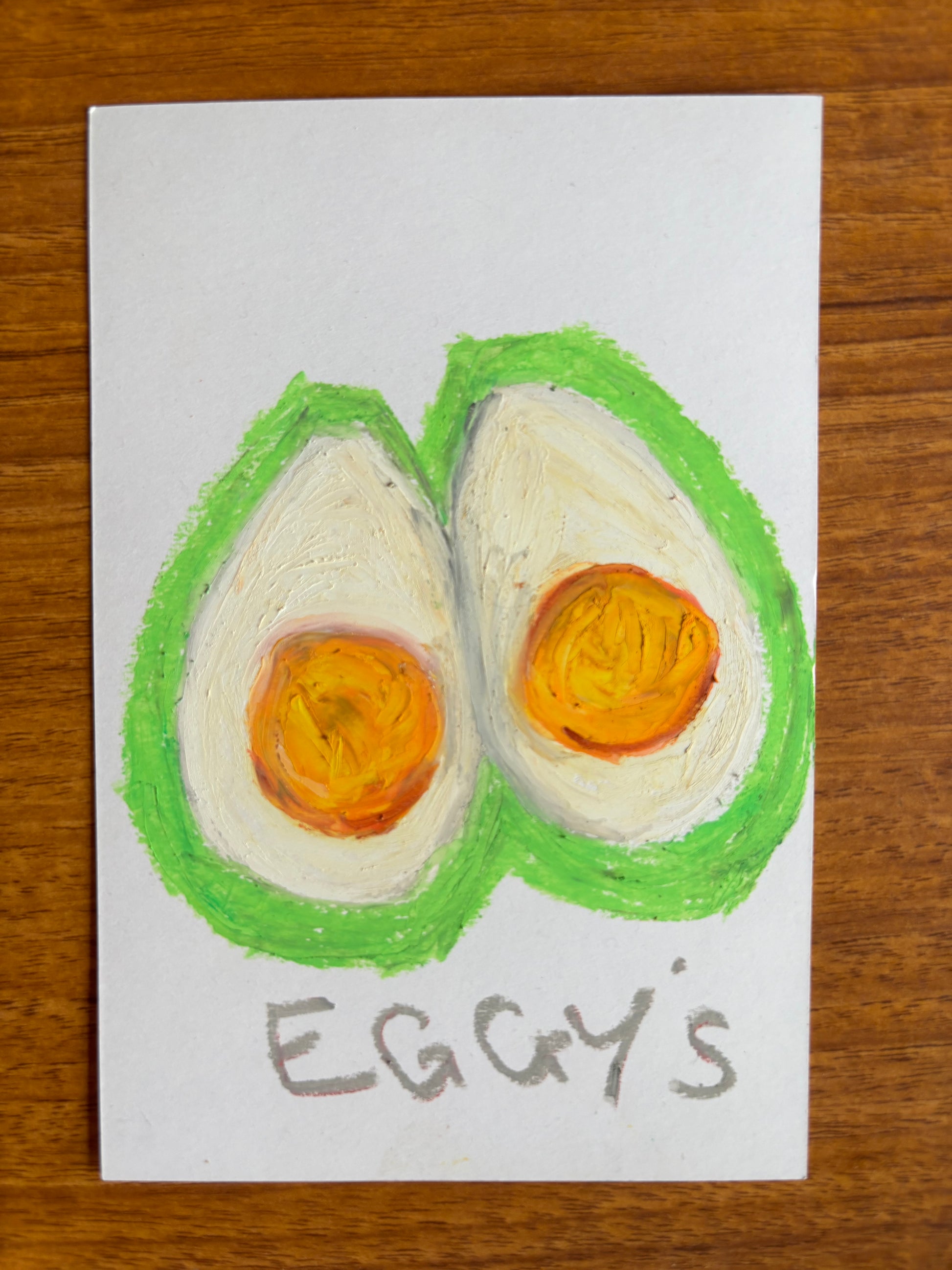 Two egg halves hand drawn on card stock with oil pastels with a green outline and the text eggy's. 
