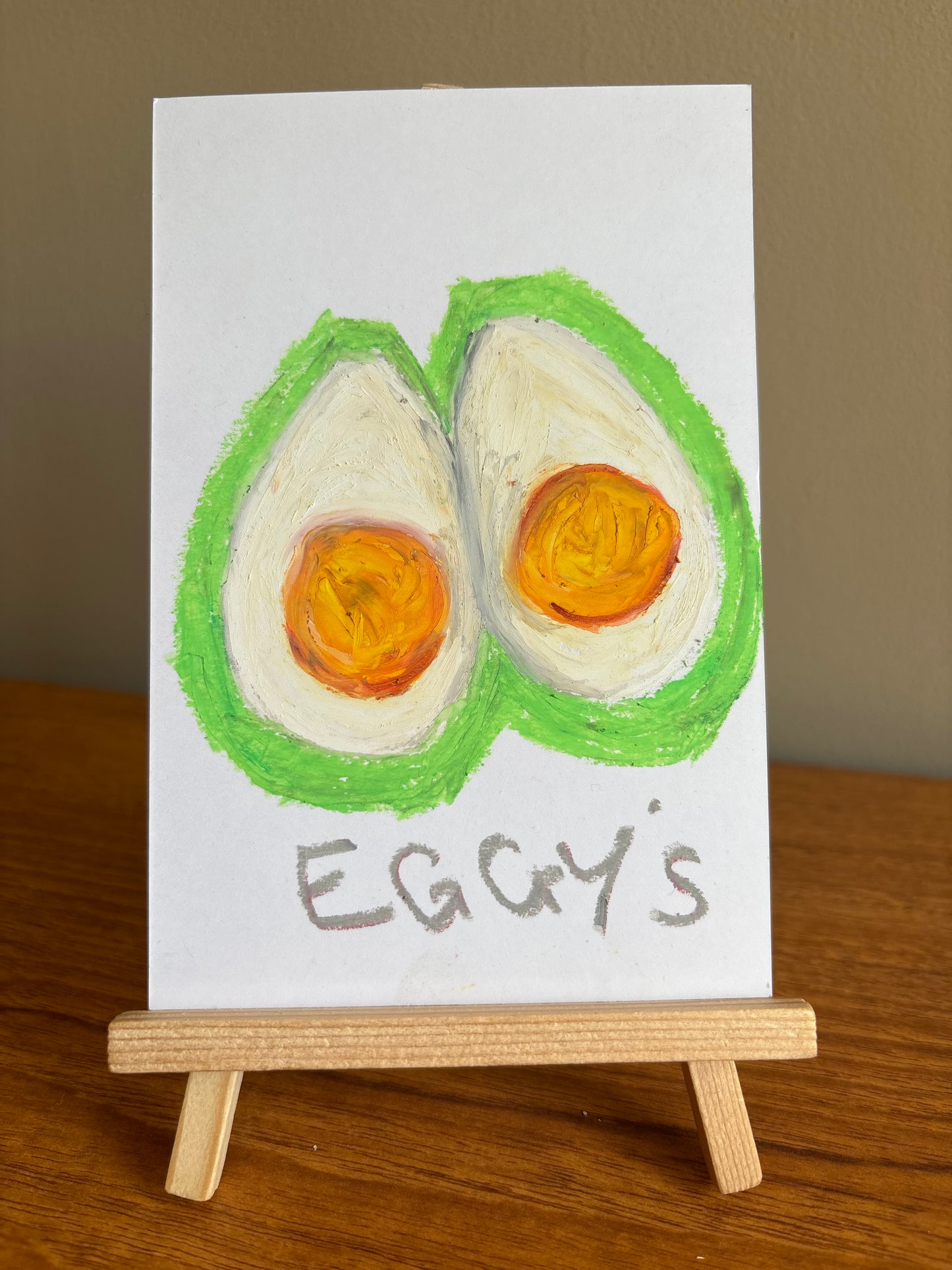 A small painted sign showing two boiled egg halves with orange yolks and the handwritten text 'EGGY'S', displayed on a wooden easel on a wooden table against a beige wall.