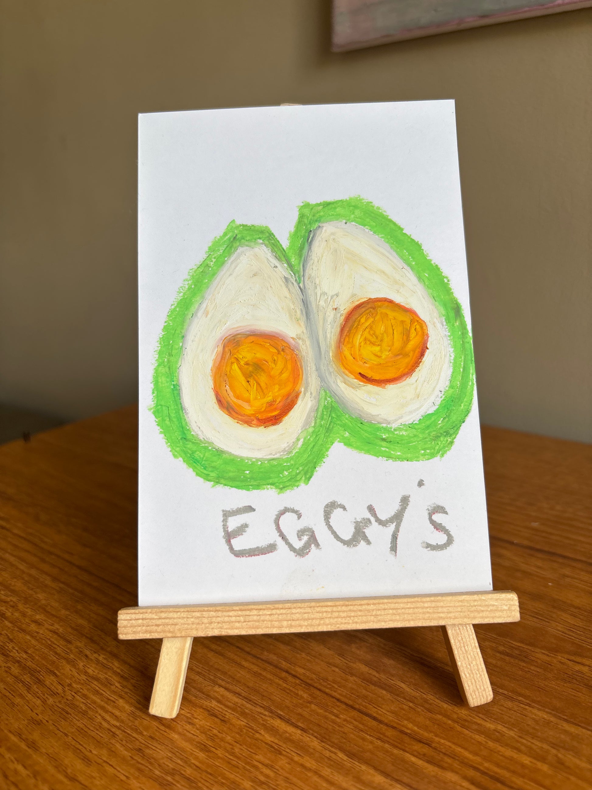A small hand-painted artwork on a white card showing two boiled egg halves with orange yolks and green outline, with the text 'EGGY'S' written below, displayed on a miniature wooden easel on a wooden table.