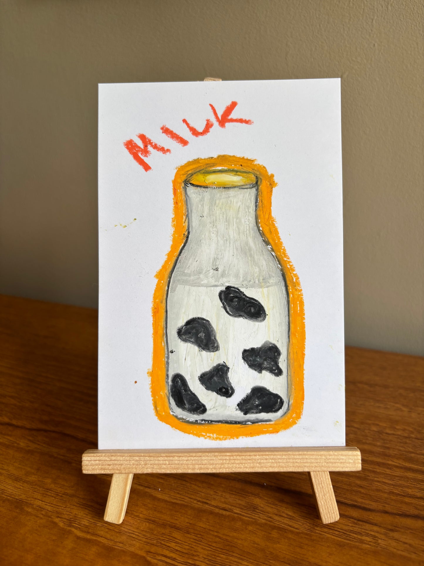 A small hand-painted artwork on a white card, showing a milk bottle with black spots and the word 'MILK' in orange above it, displayed on a wooden easel on a wooden table against a beige wall.