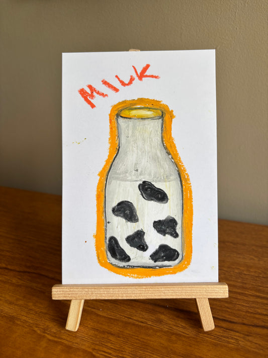A small hand-painted artwork on a white card, showing a milk bottle with black spots and the word 'MILK' in orange above it, displayed on a wooden easel on a wooden table against a beige wall.