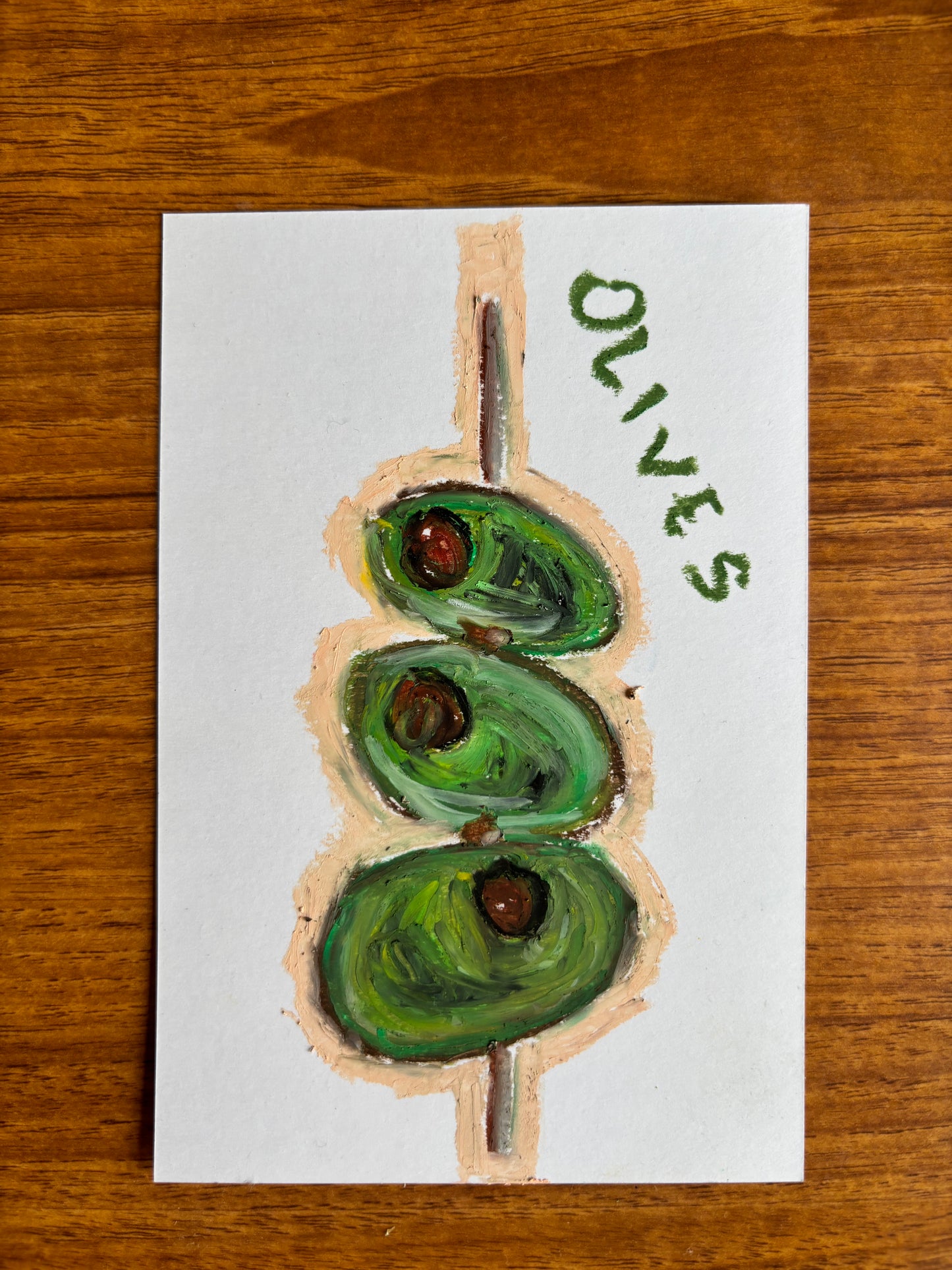 A hand-painted card featuring three green olives on a wooden skewer, with the word 'OLIVES' written in green paint on the right side, displayed on a wooden table.