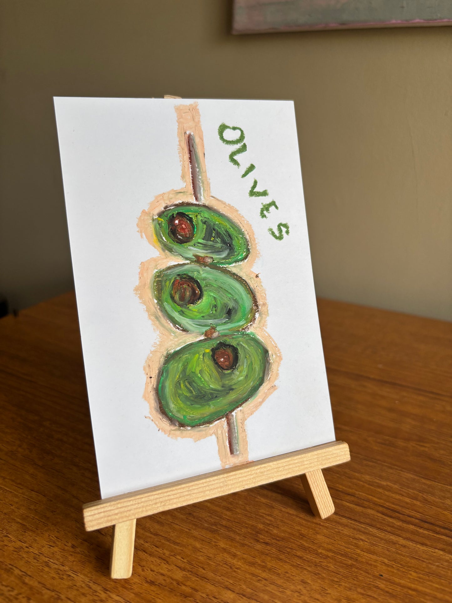 A small white card painted with three green olives on a wooden skewer and the word “OLIVES” in green, displayed on a small wooden easel on a wooden table.