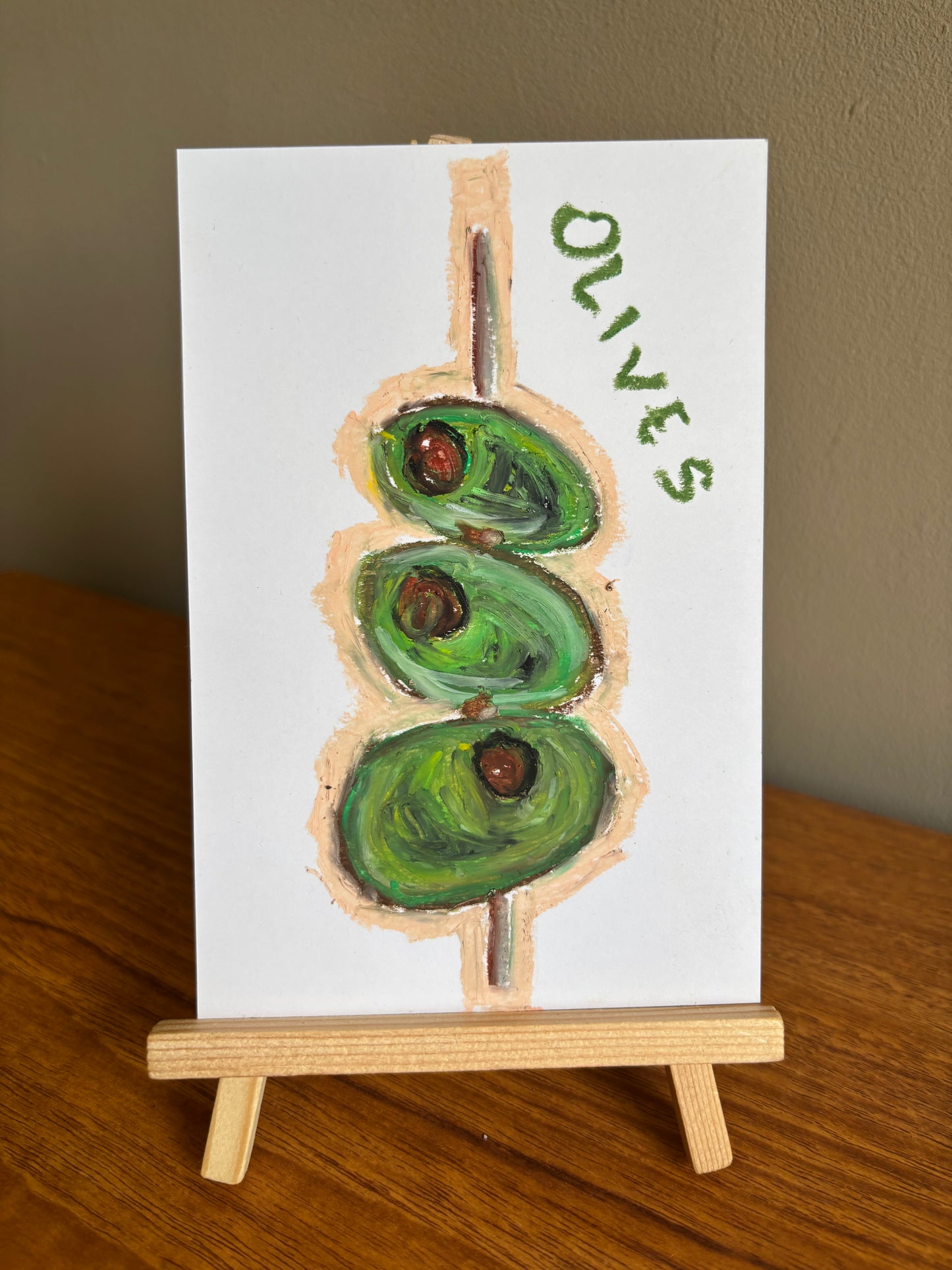 A small white art print or card showing a painted green olive on a stick with the word "OLIVES" in green, displayed on a wooden easel stand on a wooden table against a beige wall.