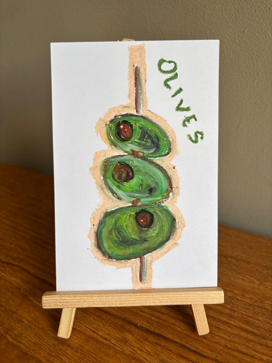A small white art print or card showing a painted green olive on a stick with the word "OLIVES" in green, displayed on a wooden easel stand on a wooden table against a beige wall.