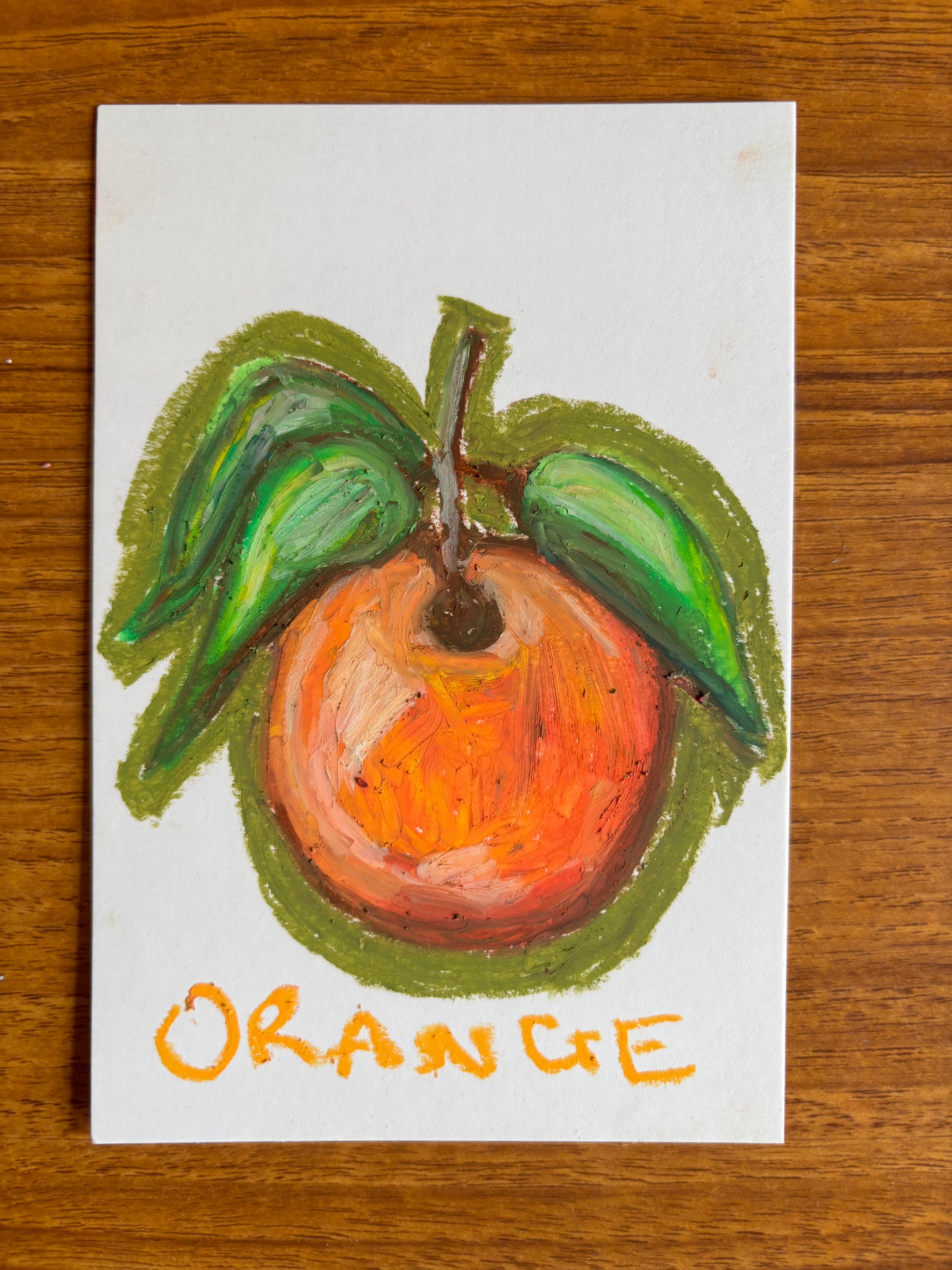 small hand drawn orange with green leaves drawn in oil pastel on a small piece of card displayed on a wooden table