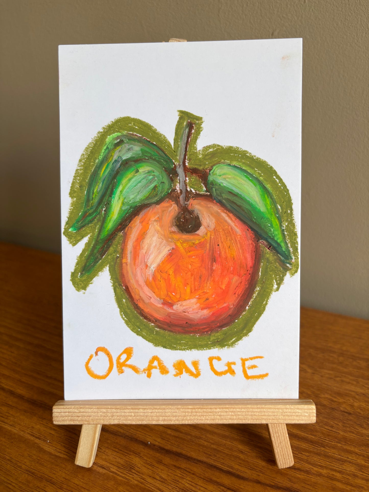 A small white art card on a wooden easel, painted with an orange fruit, green leaves, and the word 'ORANGE' in yellow-orange lettering, set on a wooden table against a beige wall.