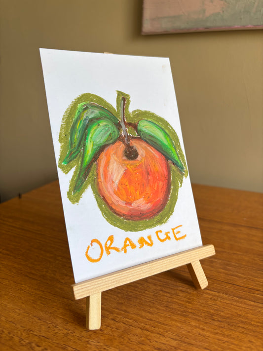 A small hand-painted pastel drawing of an orange with green leaves on a white card, displayed on a miniature wooden easel on a wooden table.