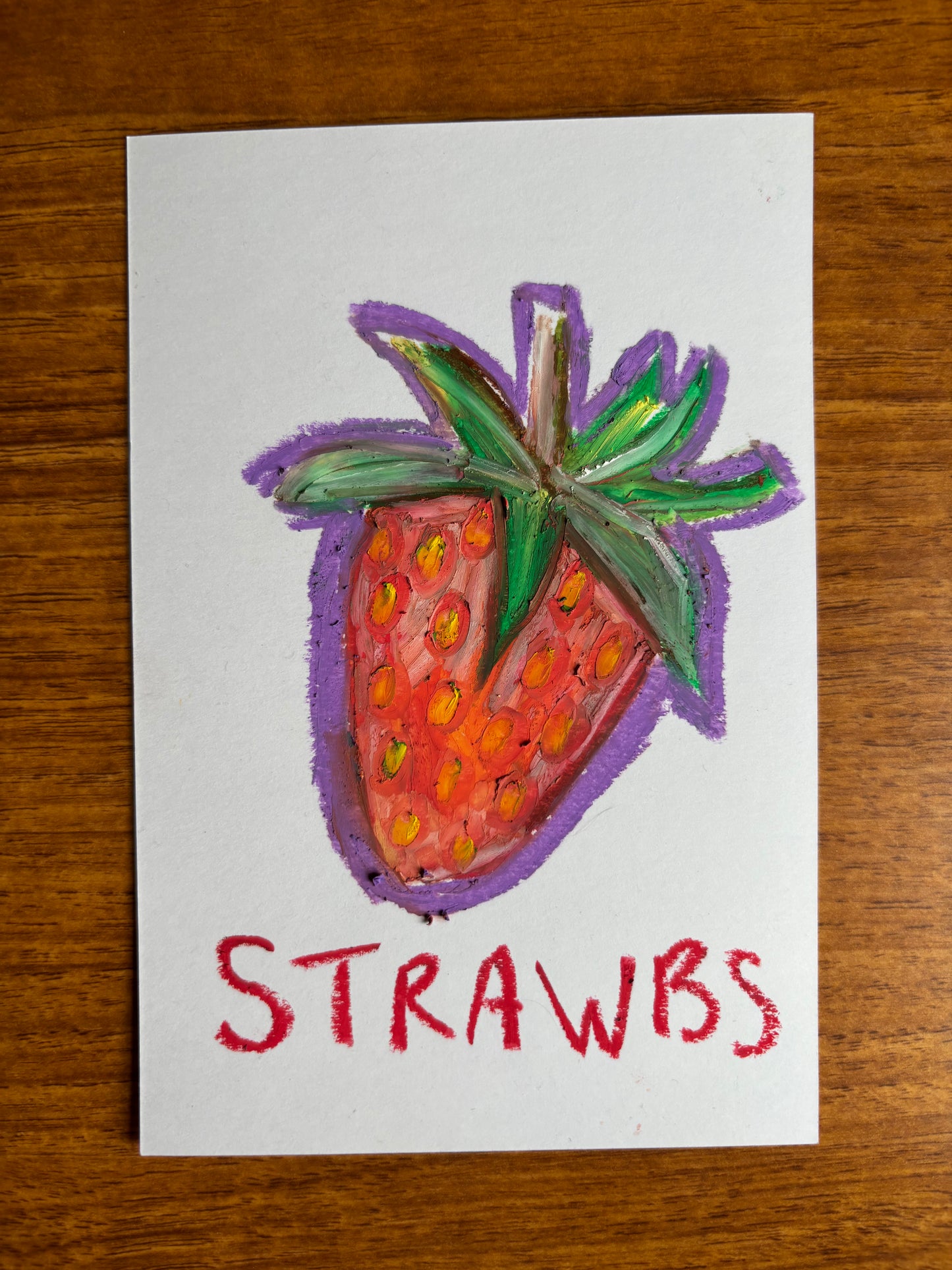 An oil pastel drawing of a strawberry on a white card, placed on a wooden table. The drawing features a red strawberry with green leaves and a purple outline, with the word 'STRAWBS' written in red below it.
