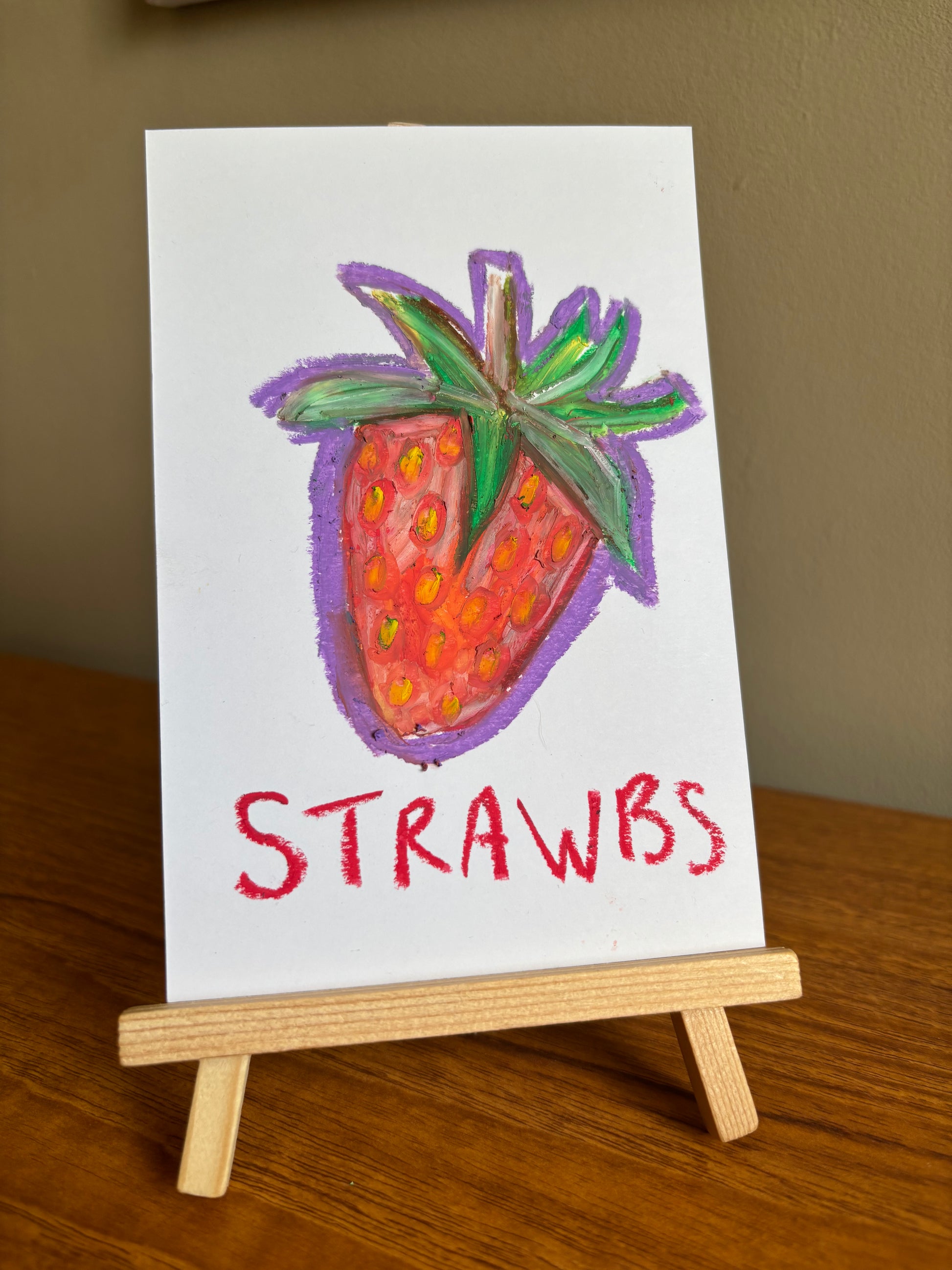 A small white card or sign on a wooden easel, featuring a colorful oil pastel drawing of a strawberry with the word “STRAWBS” written below in red.