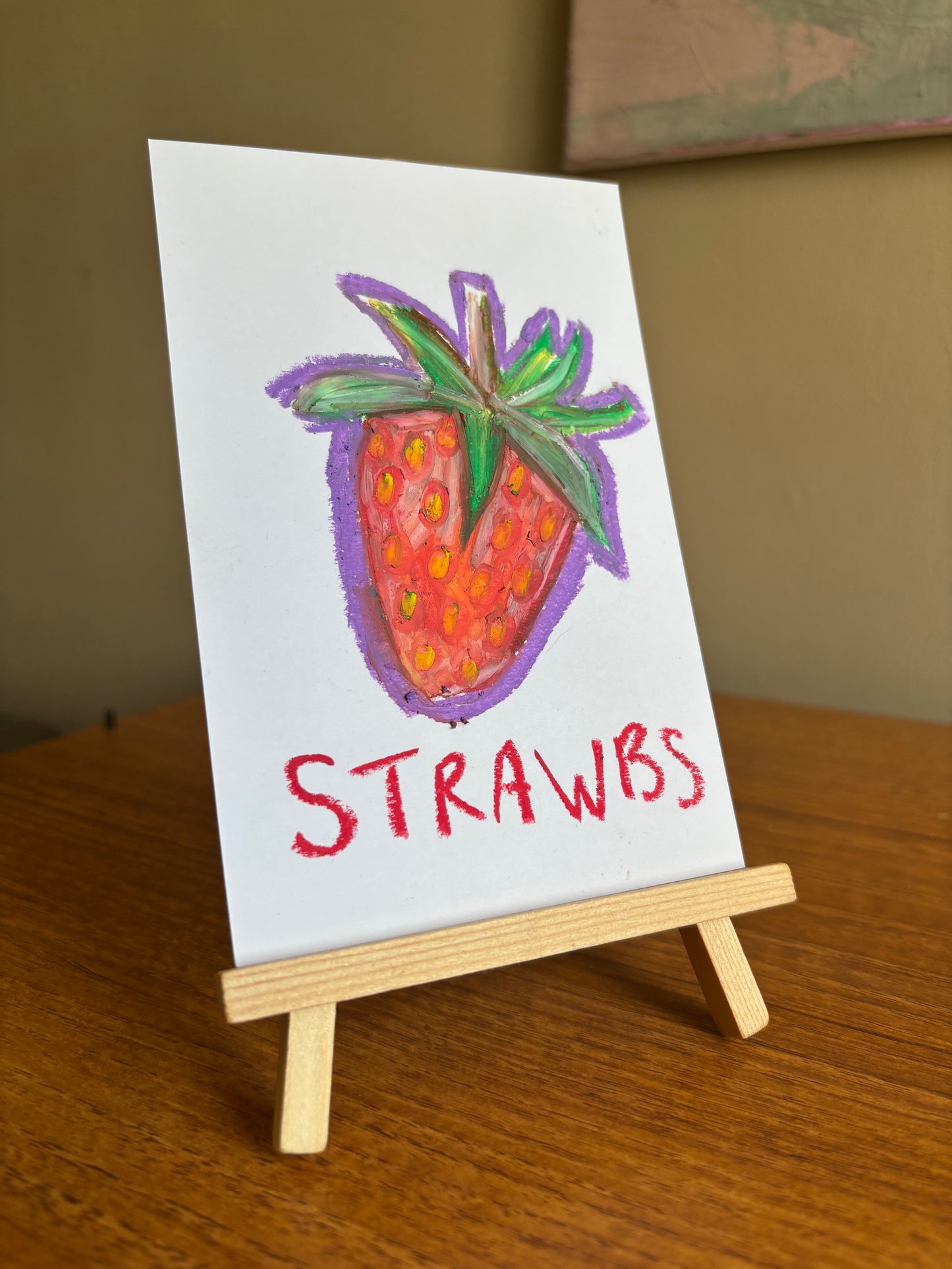 A small white card on a miniature wooden easel, featuring a hand-drawn strawberry with green leaves and the word 'STRAWBS' in red lettering, set on a wooden table.