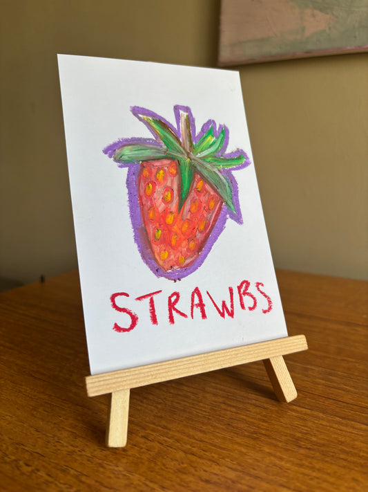 A small white card on a miniature wooden easel, featuring a hand-drawn strawberry with green leaves and the word 'STRAWBS' in red lettering, set on a wooden table.